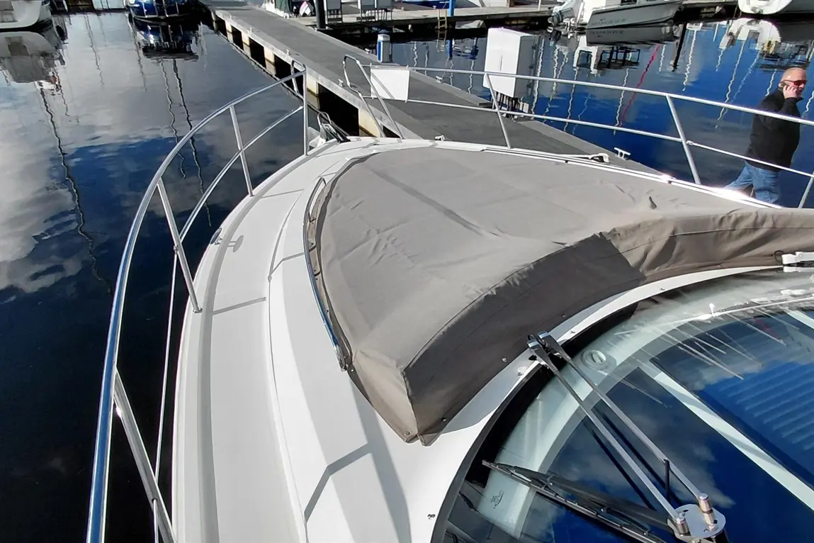 Boat deck with a gray cover and stainless steel railing, reflecting in calm water, with boats and dock in the background.